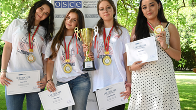 Four young women smiling, wearing medals, and holding certificates and a trophy outdoors, standing in front of a logo banner.