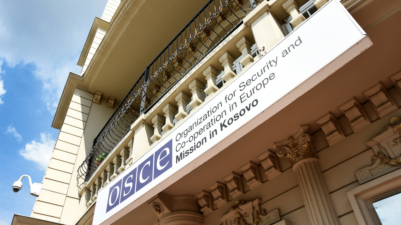 Building facade with "OSCE" logo and text, featuring ornate architectural details, seen from below against a blue sky.